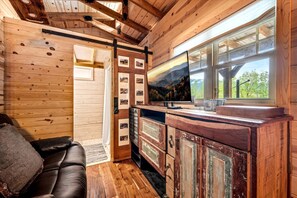 A cabin interior with a wooden ceiling, a TV on a wooden cabinet, a leather sofa, and a window with a view of trees. - Appalachian Adventure in this Tiny Cabin near Hiawassee, Ideal for Couples' Glamping (Hayesville)