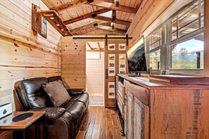 Interior de una cabaña con detalles de madera, un sofá de cuero, una mesita y una ventana con vistas al exterior.