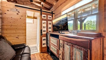 A cabin interior with a wooden ceiling, a TV on a wooden cabinet, a leather sofa, and a window with a view of trees.