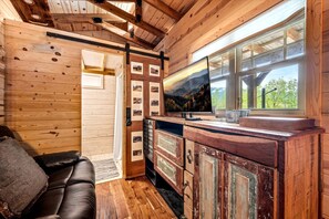 A cabin interior with a wooden ceiling, a TV on a wooden cabinet, a leather sofa, and a window with a view of trees.