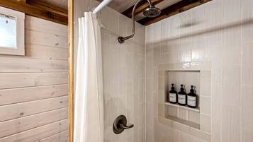 A shower with a curved showerhead, a white shower curtain, and a shelf with three bottles of shampoo, body wash, and conditioner.