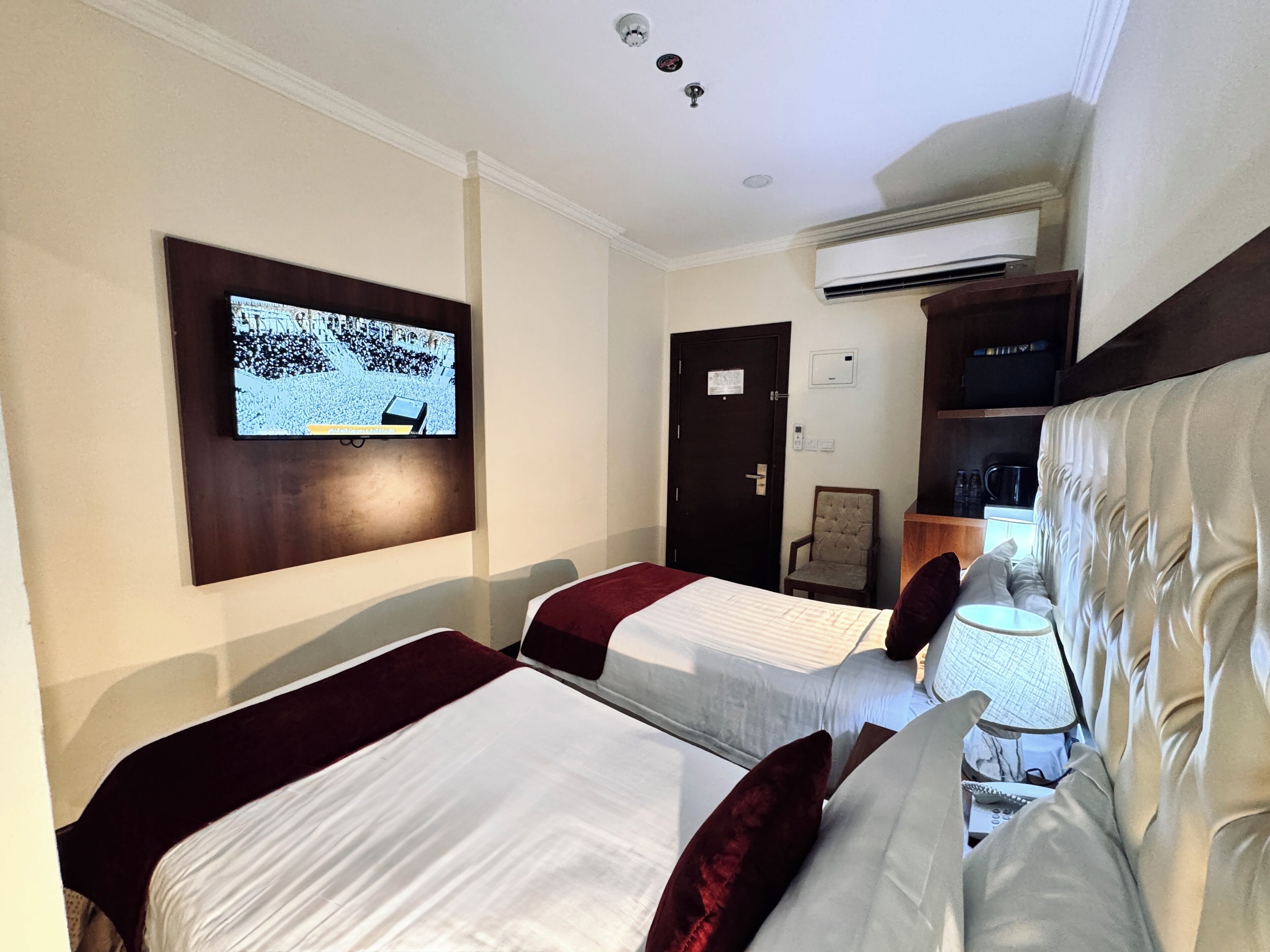 A hotel room with two beds, a television mounted on the wall, an air conditioning unit, and a small table with a lamp.