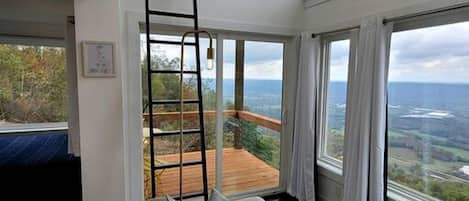 Une pièce aux hauts plafonds, dotée d’un lit en mezzanine, d’un canapé avec des coussins et d’une grande fenêtre donnant sur l’extérieur.