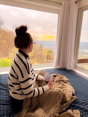 Une personne assise sur un lit, devant une fenêtre donnant sur un paysage.
