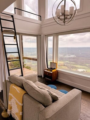 A high-ceiling room with a large window, a lofted sleeping area, and a modern chandelier.
