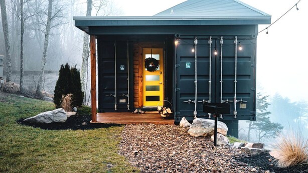 A modern building with a dark exterior, a yellow door, and a small porch area with a bench and a wreath. - Romantic Container Beautifully Design with Deck in Georgia (Rising Fawn)