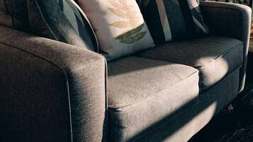 A grey sofa with patterned cushions, a wooden floor, a white door, a hanging light fixture, and a window with curtains.