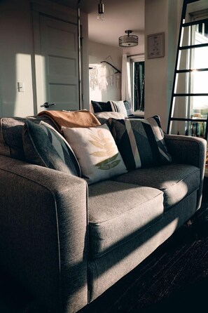 A grey sofa with patterned cushions, a wooden floor, a white door, a hanging light fixture, and a window with curtains.