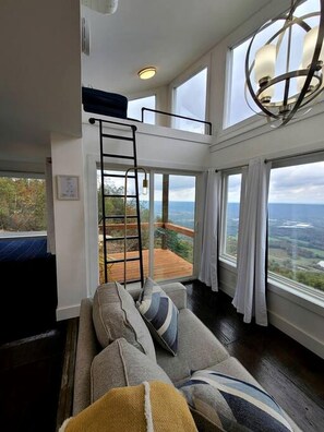 A high-ceilinged room with a loft bed, a sofa with cushions, and a large window offering a view of the outdoors. - Romantic Container Beautifully Design with Deck in Georgia (Rising Fawn)
