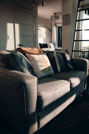 A grey sofa with patterned cushions, a wooden floor, a white door, a hanging light fixture, and a window with curtains. - Romantic Container Beautifully Design with Deck in Georgia (Rising Fawn)