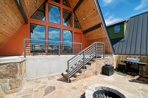 Un bâtiment moderne doté d’une terrasse en pierre, d’un espace barbecue et d’un escalier menant à un niveau supérieur avec de grandes fenêtres.