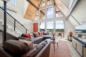 A modern living room with a large sectional sofa, a coffee table, and a TV. There is a staircase with a railing leading to an upper level.