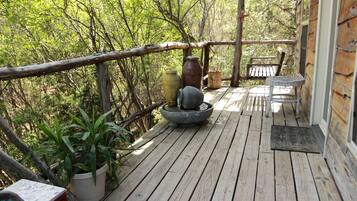 A wooden deck with a rustic railing, potted plants, a stone basin, and a wicker table.