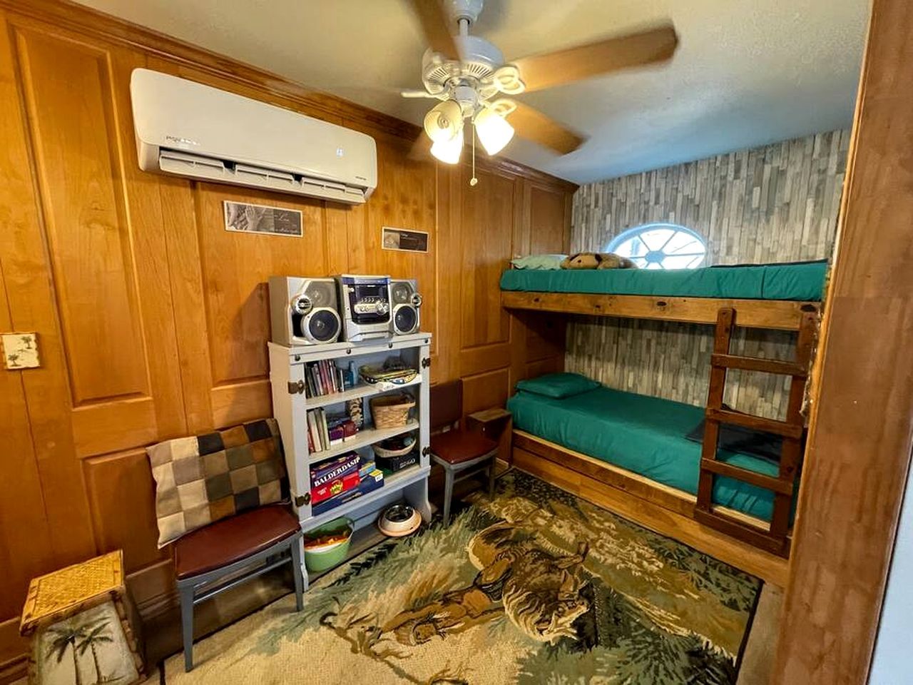 Een kleine kamer met een stapelbed, een plafondventilator, een airconditioning, een boekenplank en een klein tafeltje.