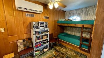 A compact room with a bunk bed, a ceiling fan, an air conditioning unit, a bookshelf, and a small table.
