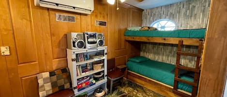 Une pièce de taille modeste, équipée d’un lit superposé, d’un ventilateur de plafond, d’un climatiseur, d’une bibliothèque et d’une petite table.