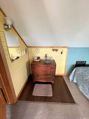 Una habitación pequeña y acogedora con un tocador de madera, un espejo, un lavabo, una cama y una alfombra pequeña.