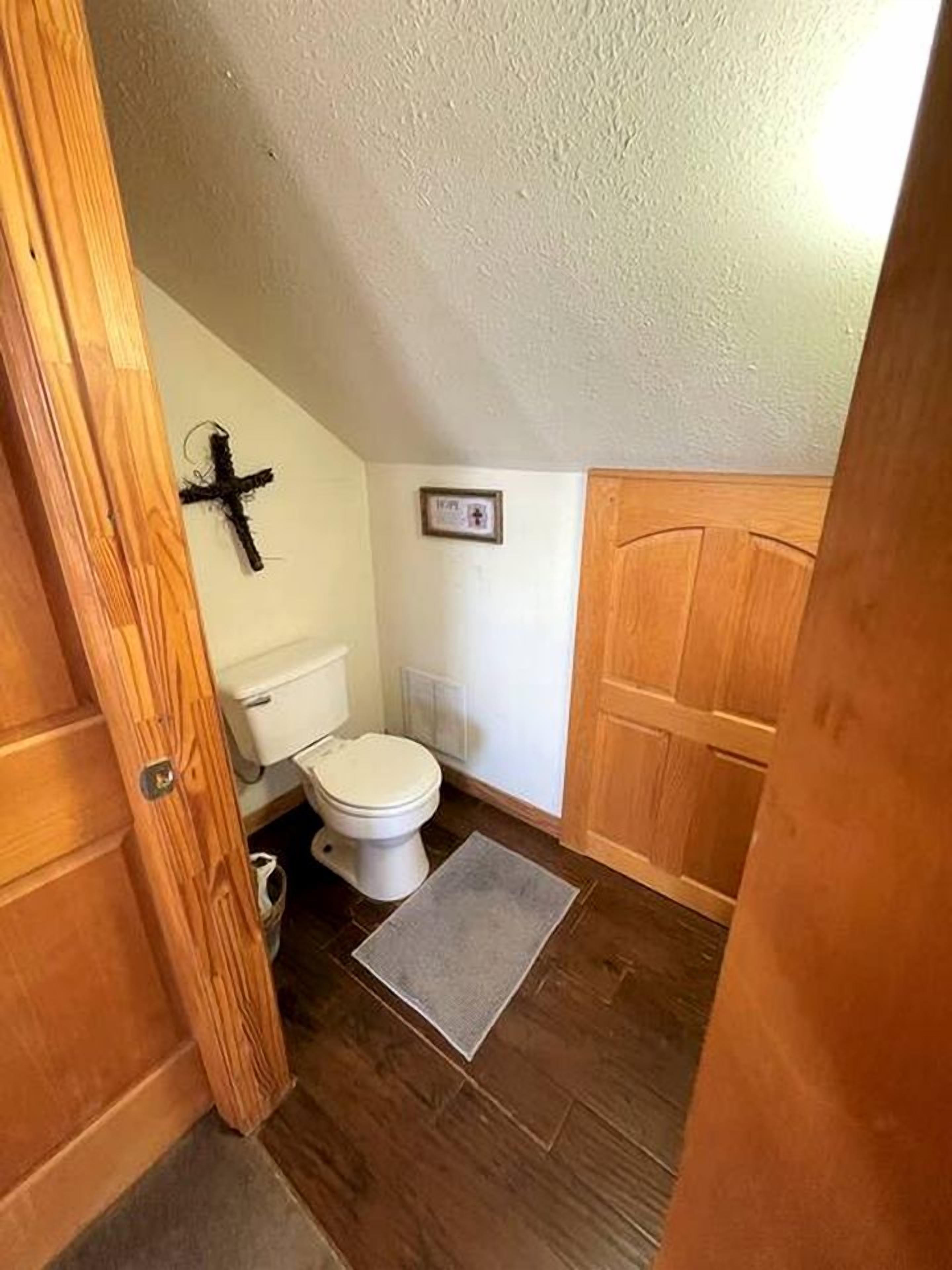 Un baño pequeño con inodoro, una puerta de madera y una cruz en la pared.