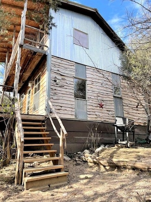 Un edificio de dos pisos con una escalera de madera que conduce a una terraza, rodeado de vegetación seca.