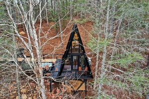A cabin with a triangular roof, surrounded by trees and a dirt ground.