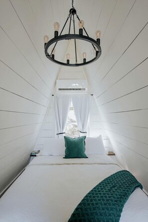 A narrow, high-ceilinged room with a bed, a chandelier, and a window with curtains.