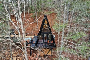 Een chalet met een puntig dak, omringd door bomen en een onverharde ondergrond.