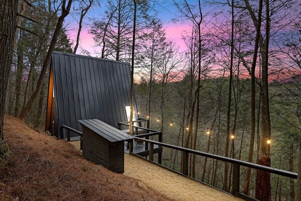 A modern cabin with a wooden deck, surrounded by trees, and a view of the sky at dusk.