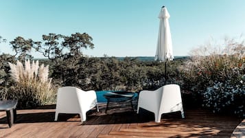 Una terraza de madera con sillas y una mesa blancas, un paraguas y vista al mar.