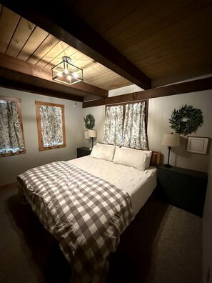 A bedroom with a checkered bedspread, wooden ceiling beams, a hanging light fixture, and a window with floral curtains.