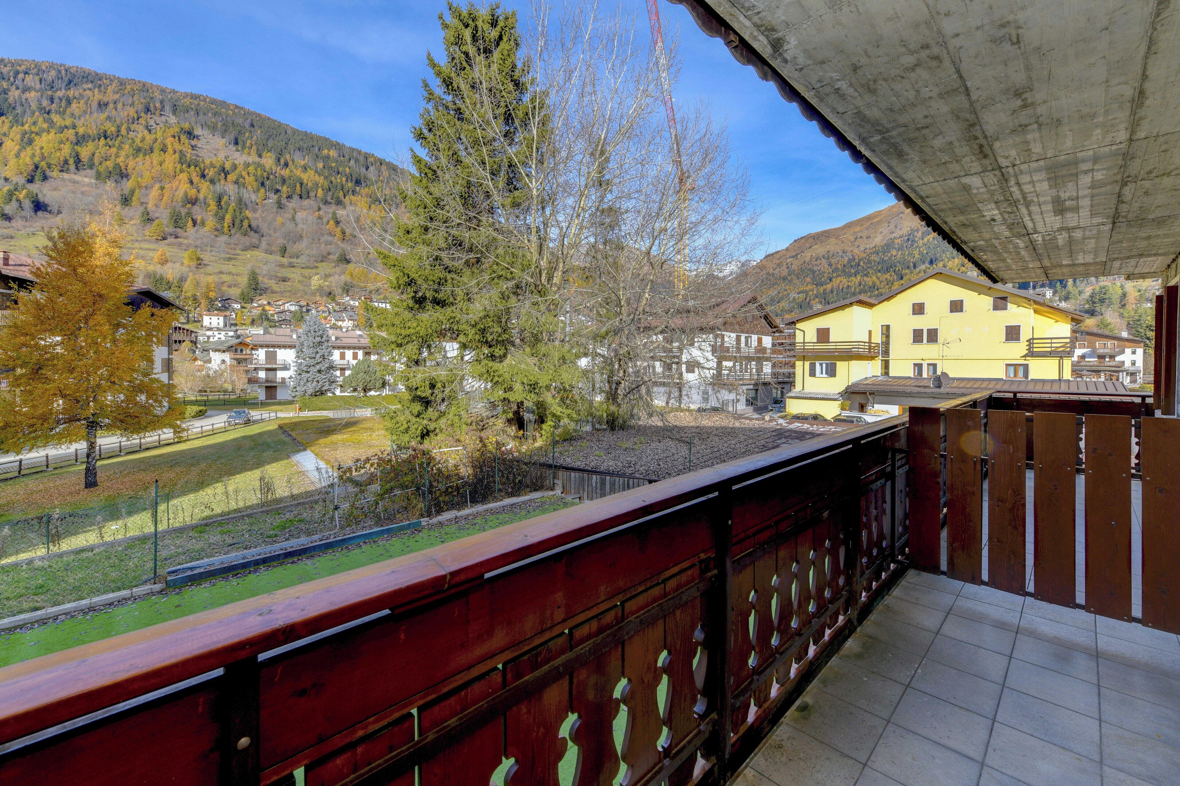 A balcony with a red railing overlooking a village with houses and a mountain in the background.