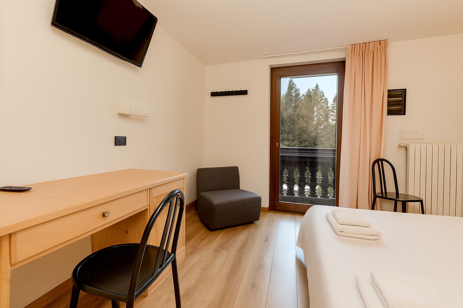Habitación de hotel con cama, escritorio, silla, televisor montado en la pared y balcón con vistas a árboles.