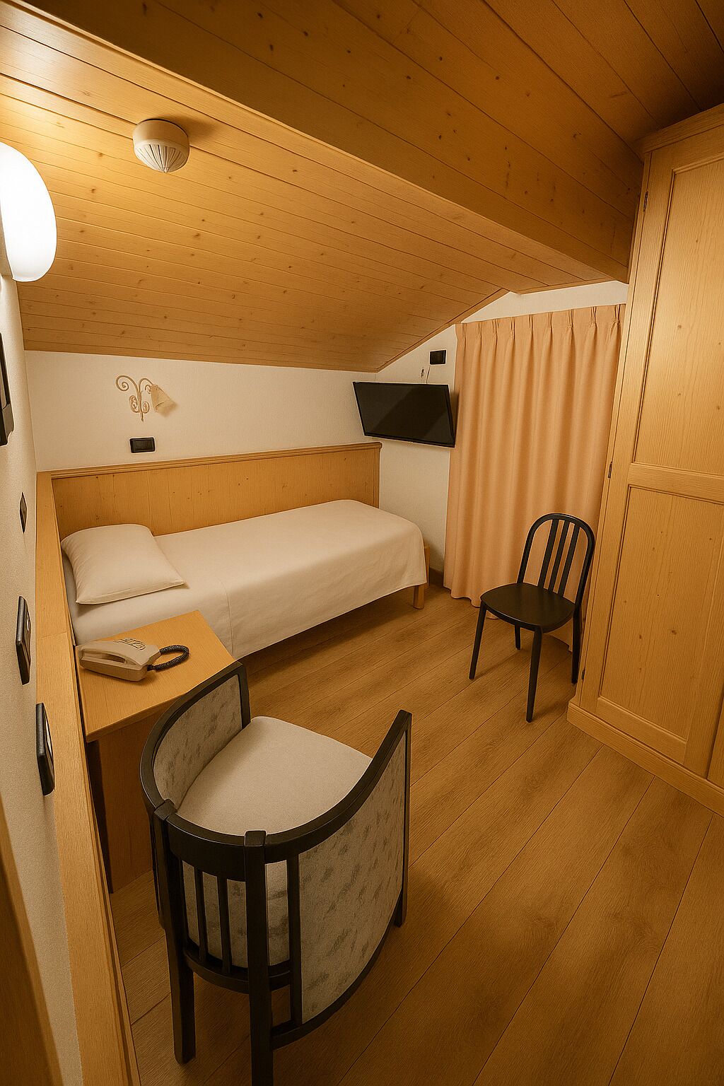 A compact room with a bed, a chair, a desk with a telephone, a television, and a ceiling-mounted light fixture.