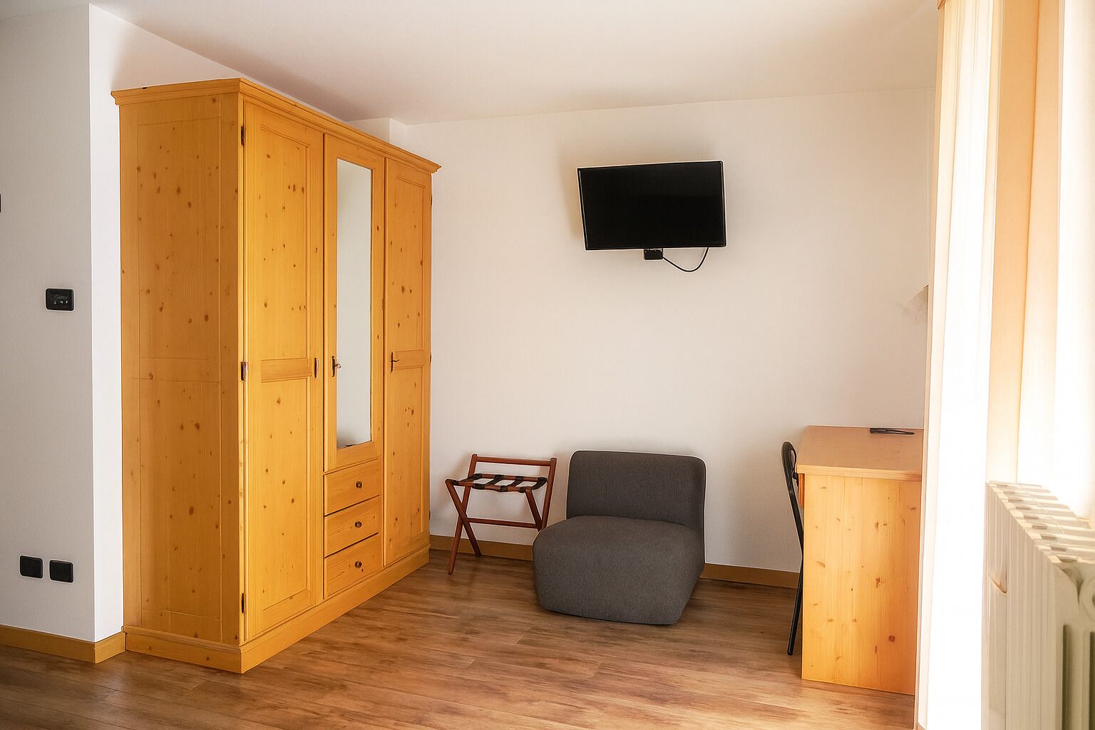 A room with a wooden wardrobe, a grey armchair, a small wooden table, and a mounted TV.
