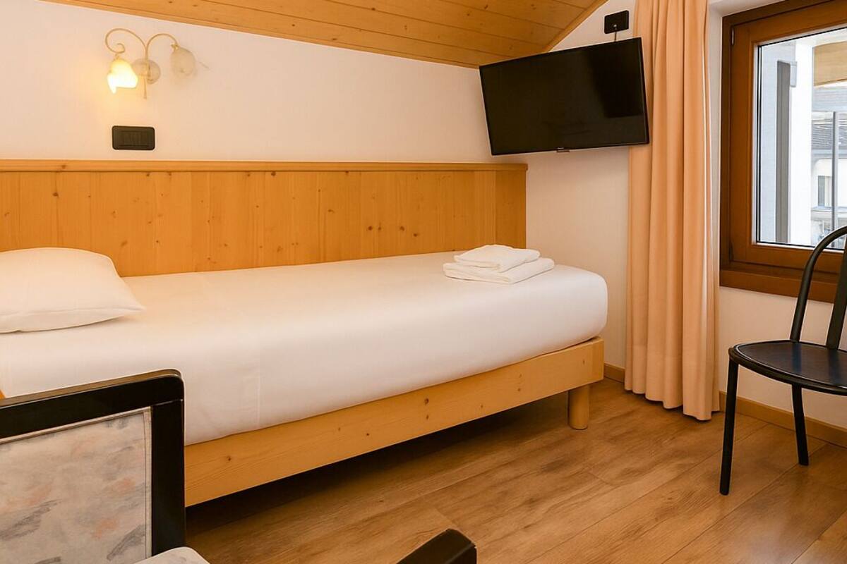 A single bed with a wooden headboard, a flat-screen TV mounted above it, and a window with curtains.