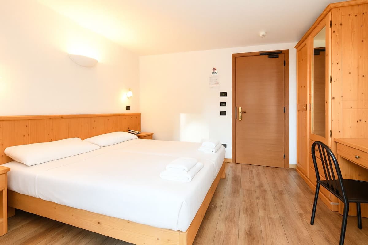 A hotel room with a large bed, wooden headboard, a desk with a chair, and a wardrobe.