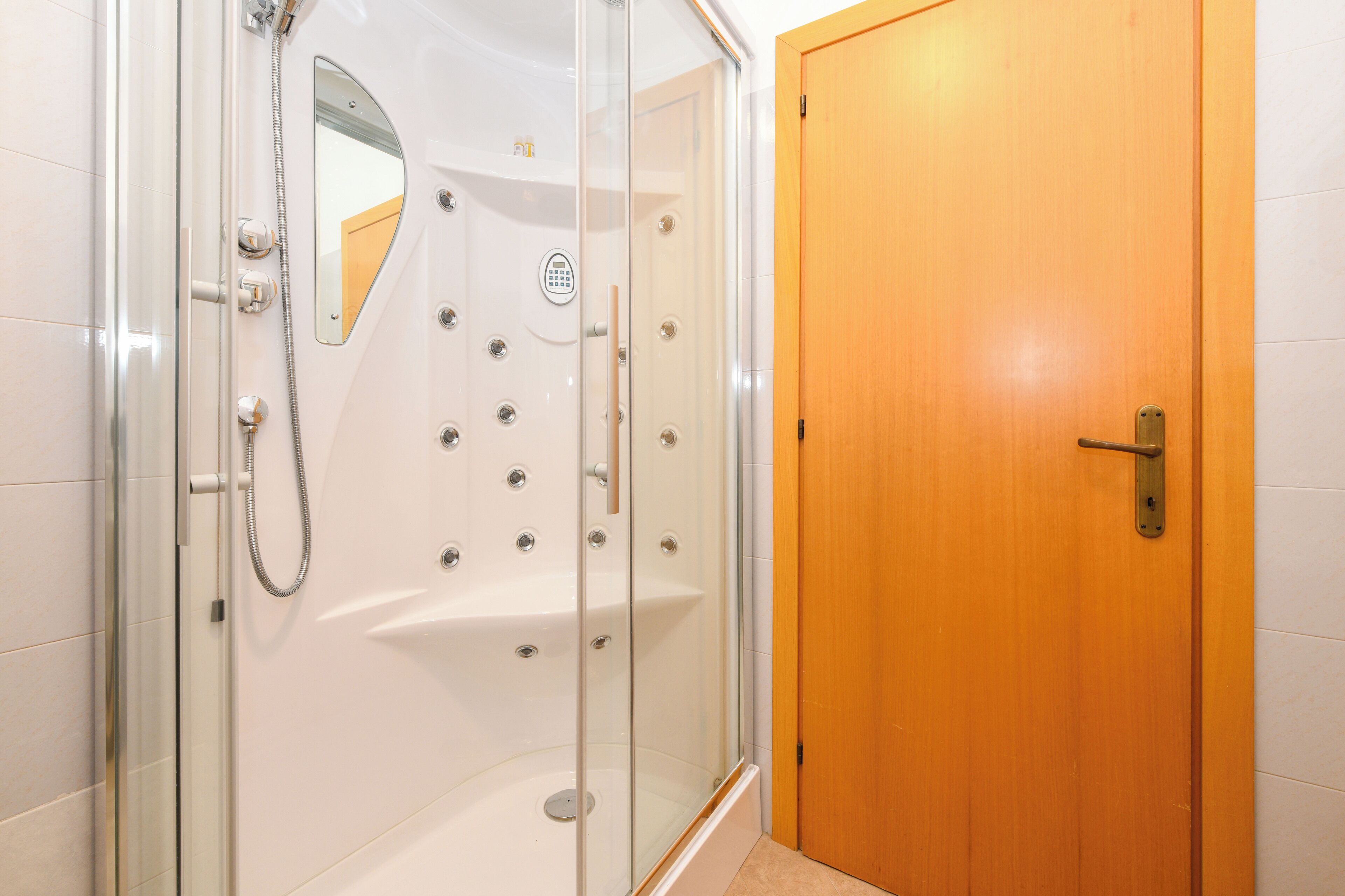 A glass-enclosed shower with multiple controls and a wooden door adjacent to it.