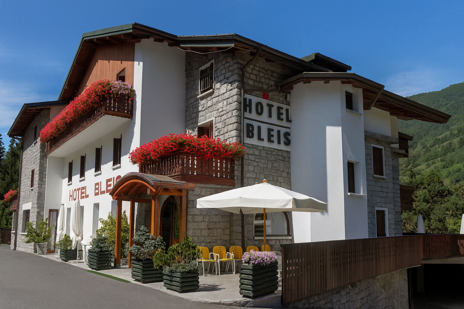 A hotel with a stone facade, balconies with red flowers, outdoor seating, and a mountain backdrop.