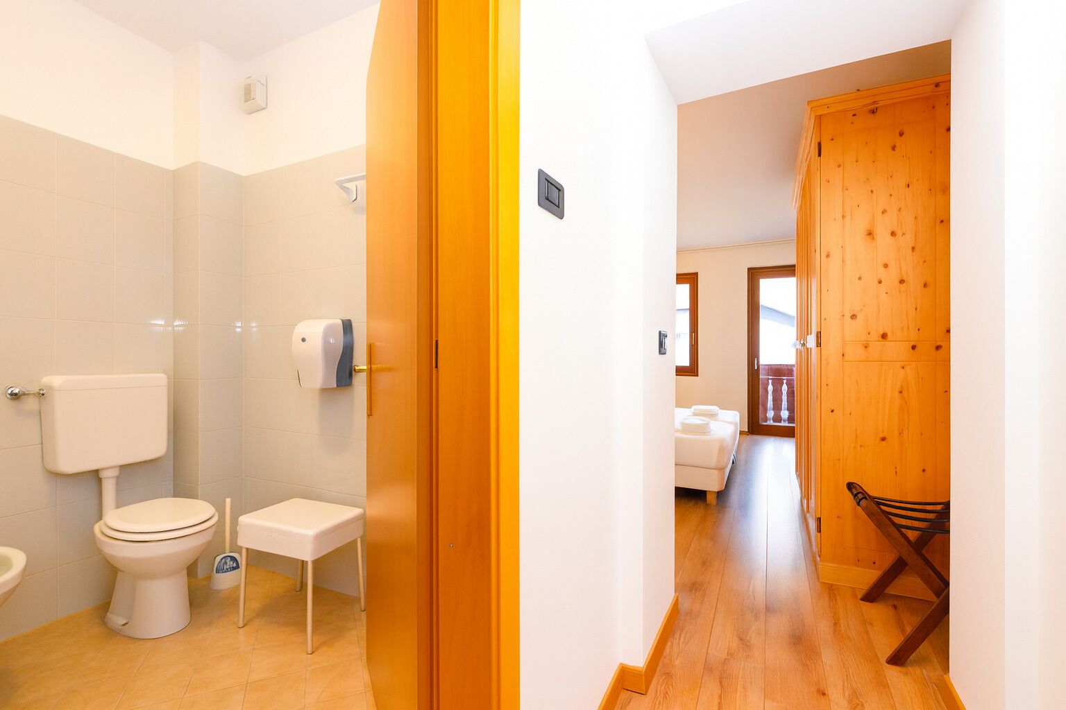 A bathroom with a toilet, a bidet, and a hand dryer. There is a wooden door leading to a bedroom with a bed and a chair.