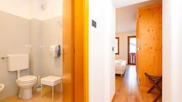 A bathroom with a toilet, a bidet, and a hand dryer. There is a wooden door leading to a bedroom with a bed and a chair.