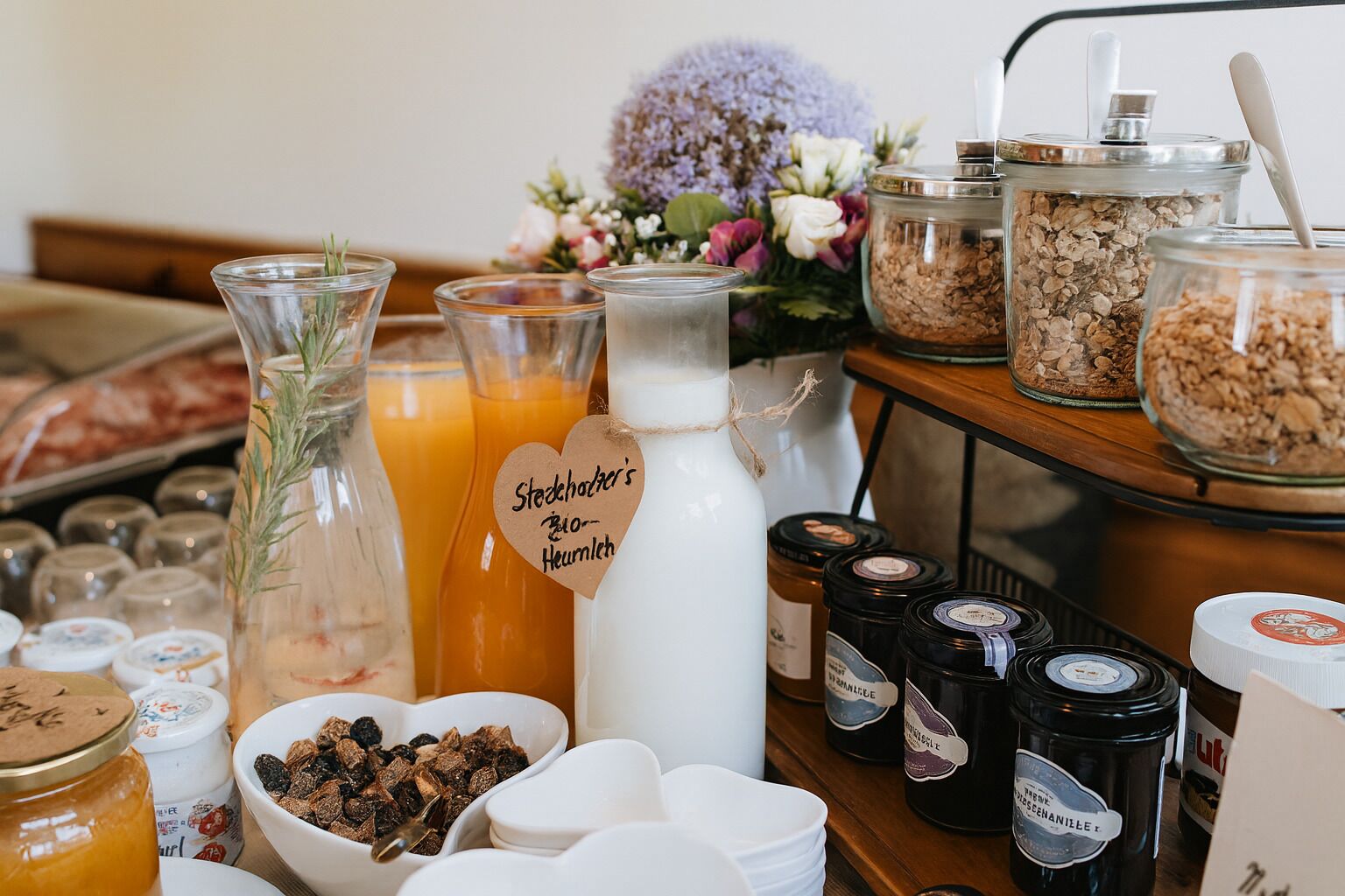 Een tafel met diverse etenswaren, waaronder potten jam, een kan melk en een schaal gedroogd fruit.