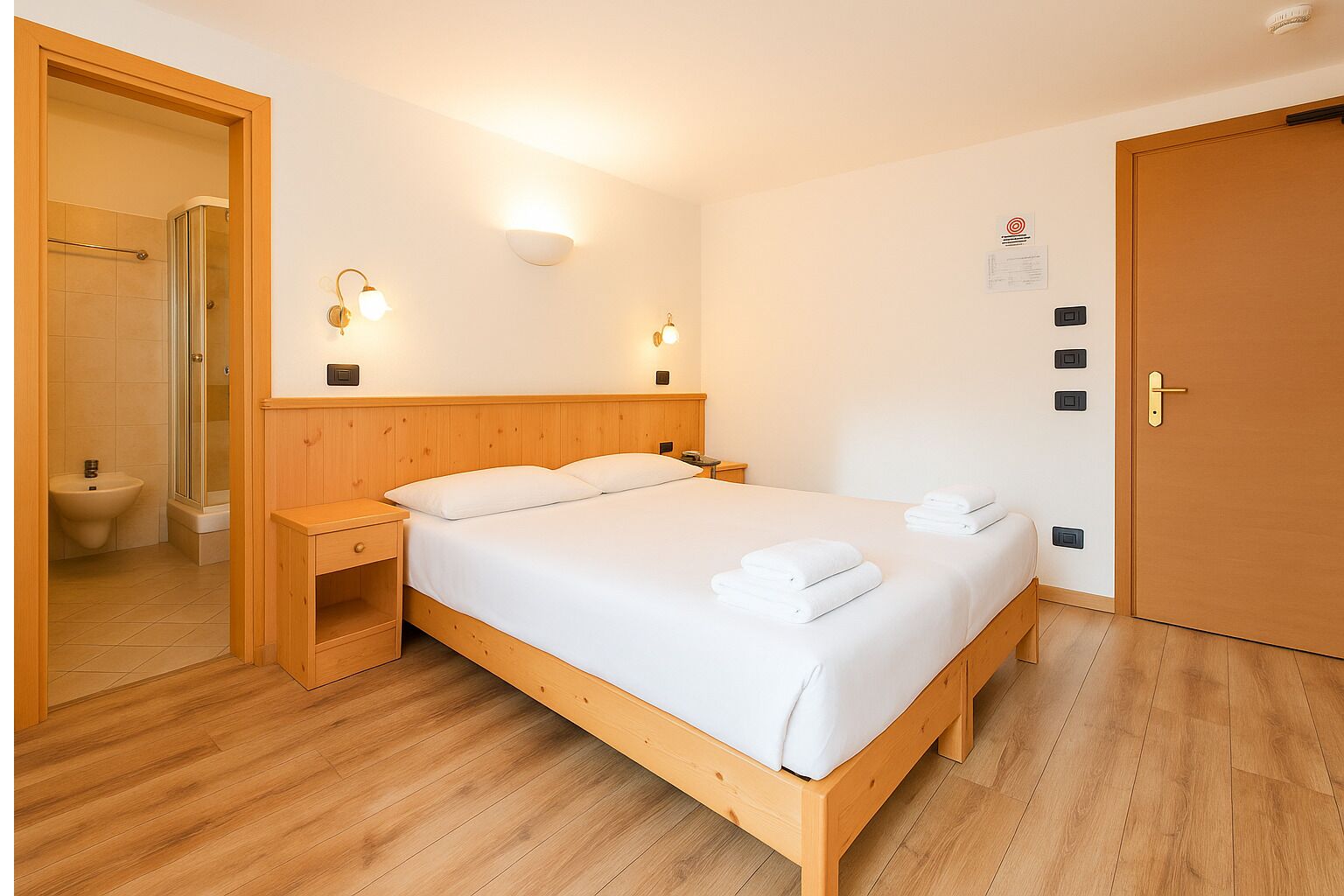 Un dormitorio con cama de madera, mesitas de noche, lámpara de pared y baño visible a través de una puerta abierta.