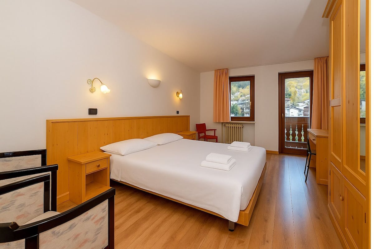 A hotel room with a large bed, wooden headboard, two bedside tables, a red chair, a desk, and a view of the outside through two windows.