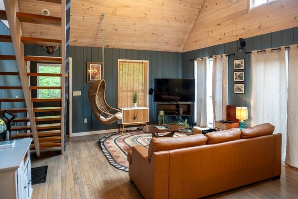 A living room with a sofa, a television, a hanging chair, a wooden staircase, and a view of the outdoors.