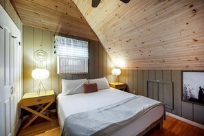 A bedroom with a sloped ceiling, a bed with white bedding, two bedside tables with lamps, a dreamcatcher on the wall, and a framed picture of llamas.