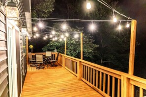 A wooden deck with a table and chairs, surrounded by a railing, and string lights overhead.
