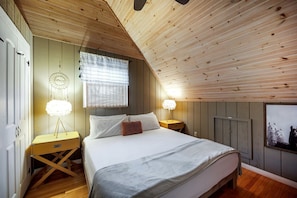 A bedroom with a sloped ceiling, a bed with white bedding, two bedside tables with lamps, a dreamcatcher on the wall, and a framed picture of llamas.