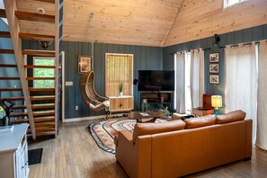 A living room with a sofa, a television, a hanging chair, a wooden staircase, and a view of the outdoors.