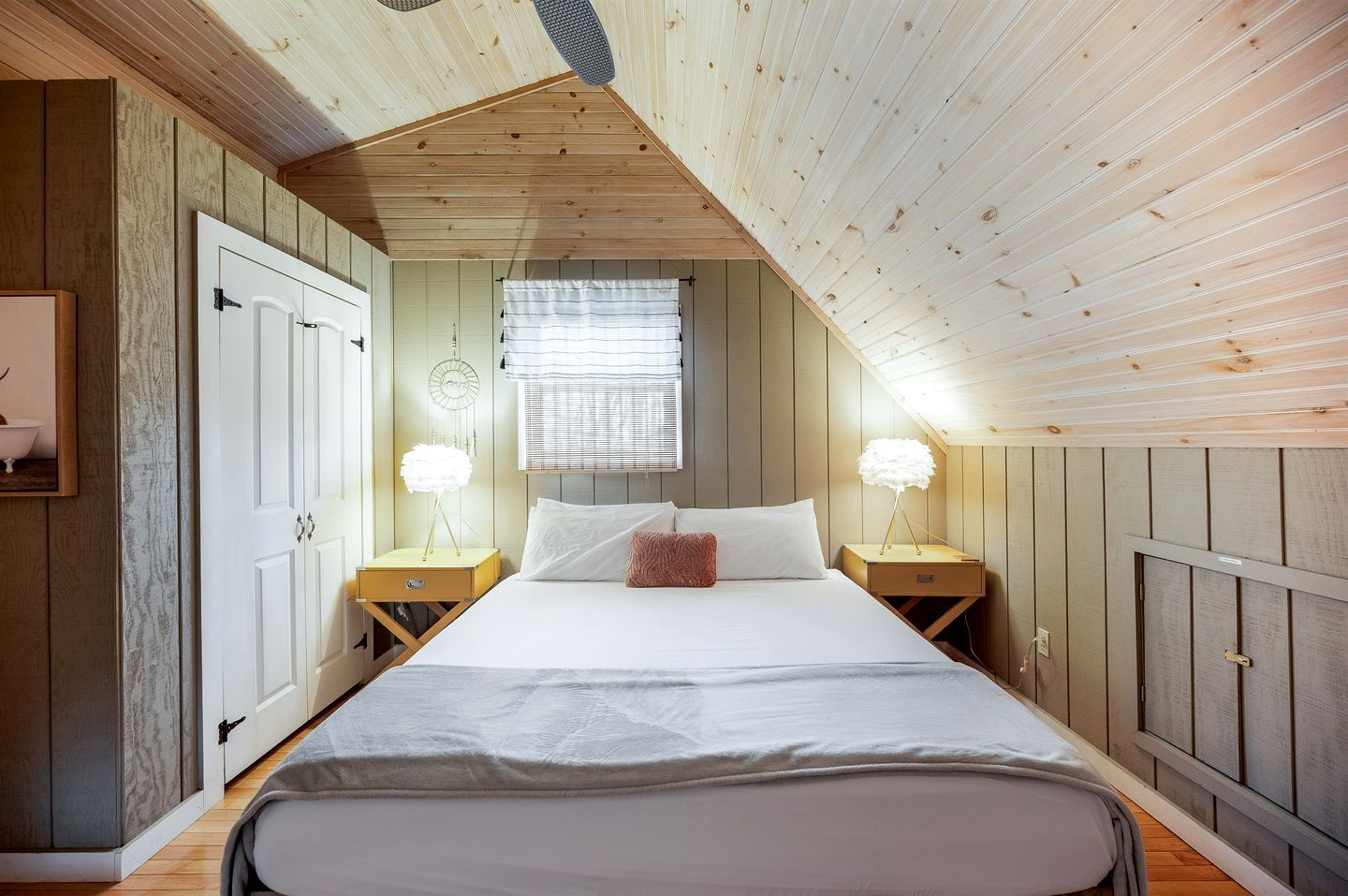 Quarto com teto inclinado, cama com roupa de cama branca, duas luminárias de cabeceira e uma janela com persianas.