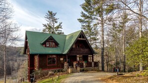 A rustic cabin with a green roof, wooden exterior, and a gravel driveway leading to a wooden porch.
