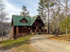 A rustic cabin with a green roof, wooden exterior, and a gravel driveway leading to a wooden porch.